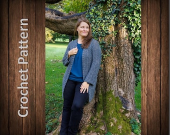 May include: A woman wearing a gray crocheted cardigan with long sleeves and a blue shirt. She is standing in front of a tree with green leaves and moss.