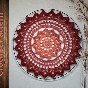 May include: A handcrafted crochet pattern in shades of red and orange, displayed on a black metal ring. The text "Crochet Pattern" is visible on the left side. The design features a central starburst pattern and a decorative outer edge.