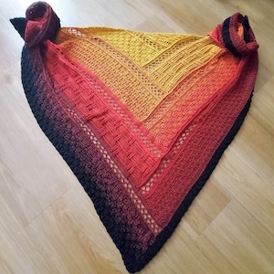 Puede incluir: Un chal de ganchillo con un degradado de colores desde el negro hasta el naranja y el amarillo. El chal tiene un patr&oacute;n de encaje y un borde negro.