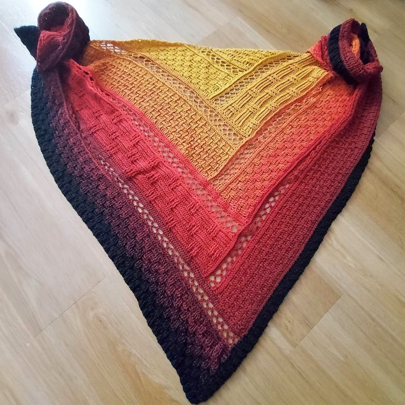 Puede incluir: Un chal de ganchillo con un degradado de colores desde el negro hasta el naranja y el amarillo. El chal tiene un patr&oacute;n de encaje y un borde negro.