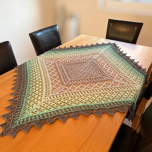 Peut inclure: Un châle carré au crochet fait à la main, dans un dégradé de couleurs, d'une bordure gris foncé à un centre vert clair et beige. Le châle est présenté sur une table en bois, mettant en valeur son design ajouré et ses bords festonnés.
