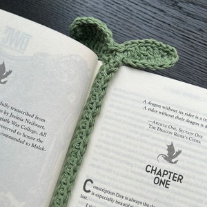 Peut inclure: Un marque-page en crochet vert sauge en forme de feuille, placé dans un livre ouvert. Le marque-page est fait d'un fil doux et a un aspect texturé. Les pages du livre sont visibles, avec du texte et les mots "CHAPTER ONE".