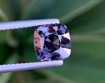 May include: A dark purple gemstone with a square cushion cut, held by tweezers against a green leafy background.