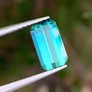 Op de afbeelding: Een teal blauwe edelsteen met een rechthoekige vorm, vastgehouden in een pincet tegen een wazige groene achtergrond.