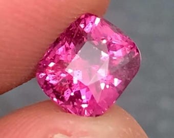 May include: A vivid, cushion-cut pink gemstone held between fingers. The gem has a square shape with rounded corners and facets that reflect light. The colour is a bright, saturated pink, and the gem appears to be of high quality.
