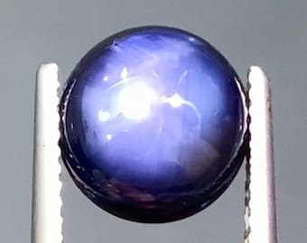 May include: A round, polished, dark blue gemstone with a star-like effect. The gem is held by silver tweezers against a gray background. The star effect is caused by light reflecting off inclusions within the stone.