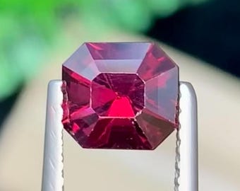 May include: A faceted, deep red gemstone held by silver tweezers. The gem has an octagonal shape with sharp, defined edges. The background is blurred, with green and neutral tones.