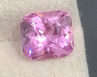 May include: A cushion-cut, faceted pink gemstone. The gem is a vibrant shade of pink, with visible internal facets that catch and reflect light. The stone is set against a white background, highlighting its color and clarity. The gemstone is likely for jewelry.