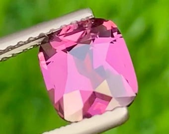 May include: A faceted, rectangular pink gemstone is held by a pair of tweezers against a blurred green background. The gem has multiple facets that reflect light, creating a sparkling effect. The tweezers are silver-colored.