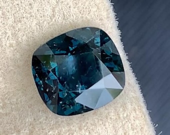 May include: A dark blue gemstone with a cushion cut. The gemstone is on a white background.