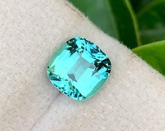 May include: A cushion-cut teal gemstone with a light blue hue. The gemstone is set on a white background.