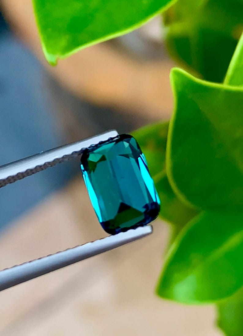 May include: A vibrant green gemstone with a rectangular cut, held by tweezers against a green leafy background.