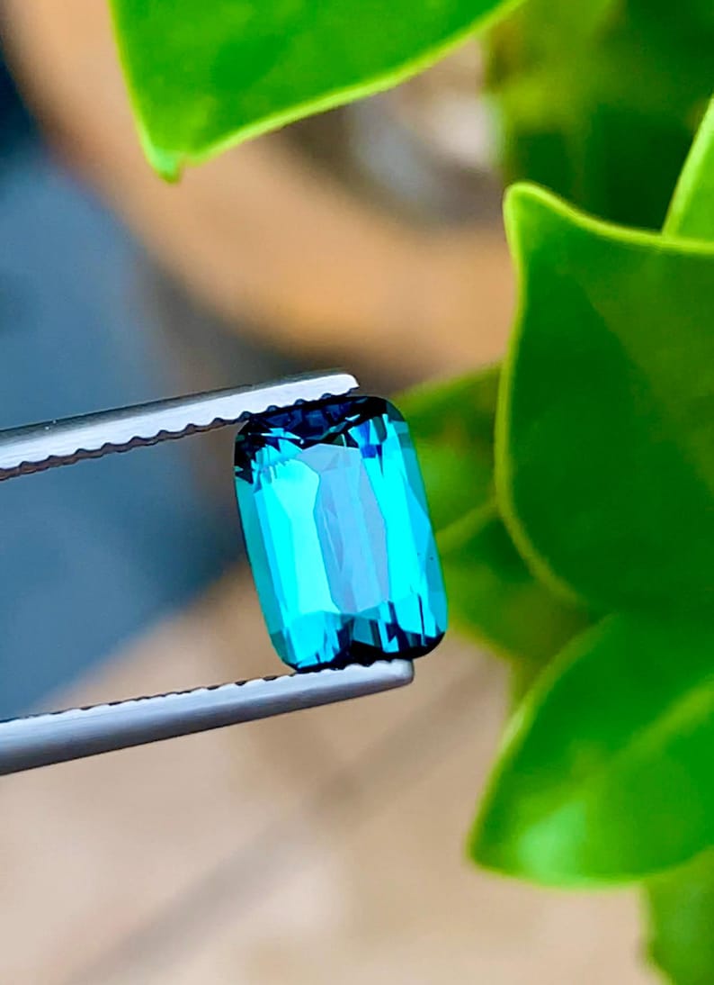 May include: A vibrant blue-green gemstone with a rectangular cut, held in tweezers against a green leafy background.