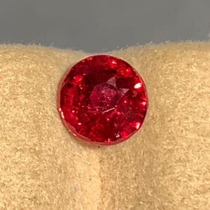 May include: A round, faceted gemstone with a deep, vibrant red color. The stone features a brilliant cut with multiple triangular and kite-shaped facets that reflect light, creating a sparkling appearance across its circular surface.
