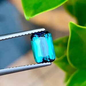 May include: A vibrant teal green gemstone with a rectangular cut, held in tweezers against a green leafy background.