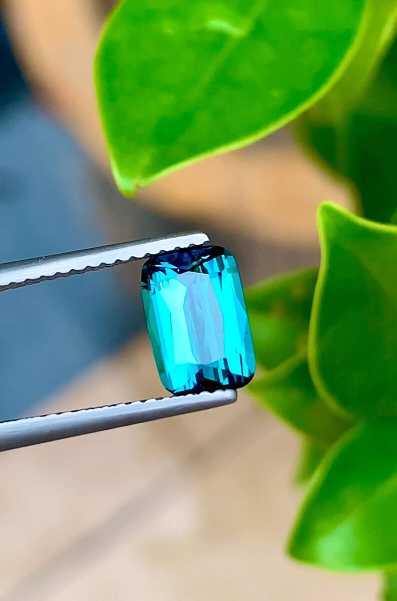 May include: A vibrant teal green gemstone with a rectangular cut, held in tweezers against a green leafy background.