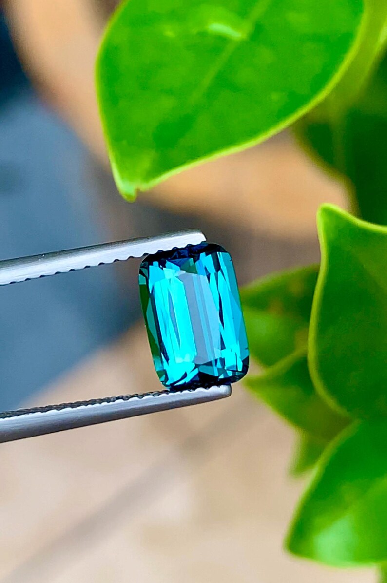 May include: A vibrant green gemstone with a rectangular cut, held in tweezers against a green leafy background.