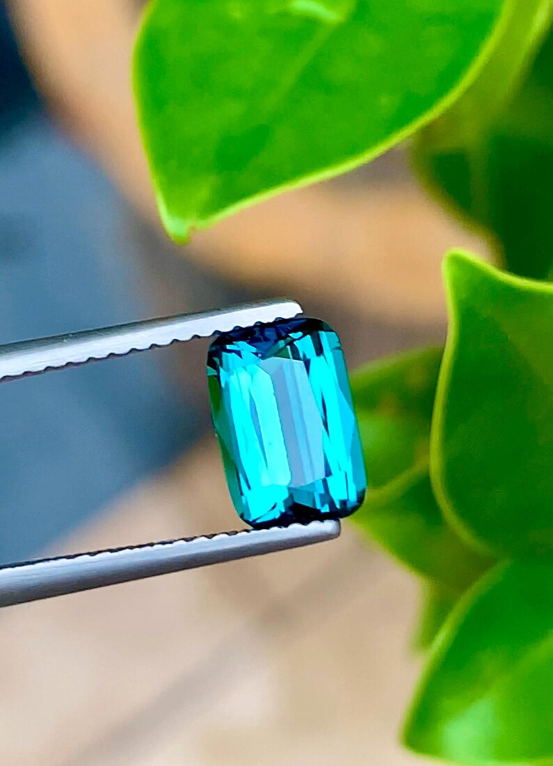 May include: A vibrant blue-green gemstone with a rectangular shape, held in tweezers against a green leafy background.