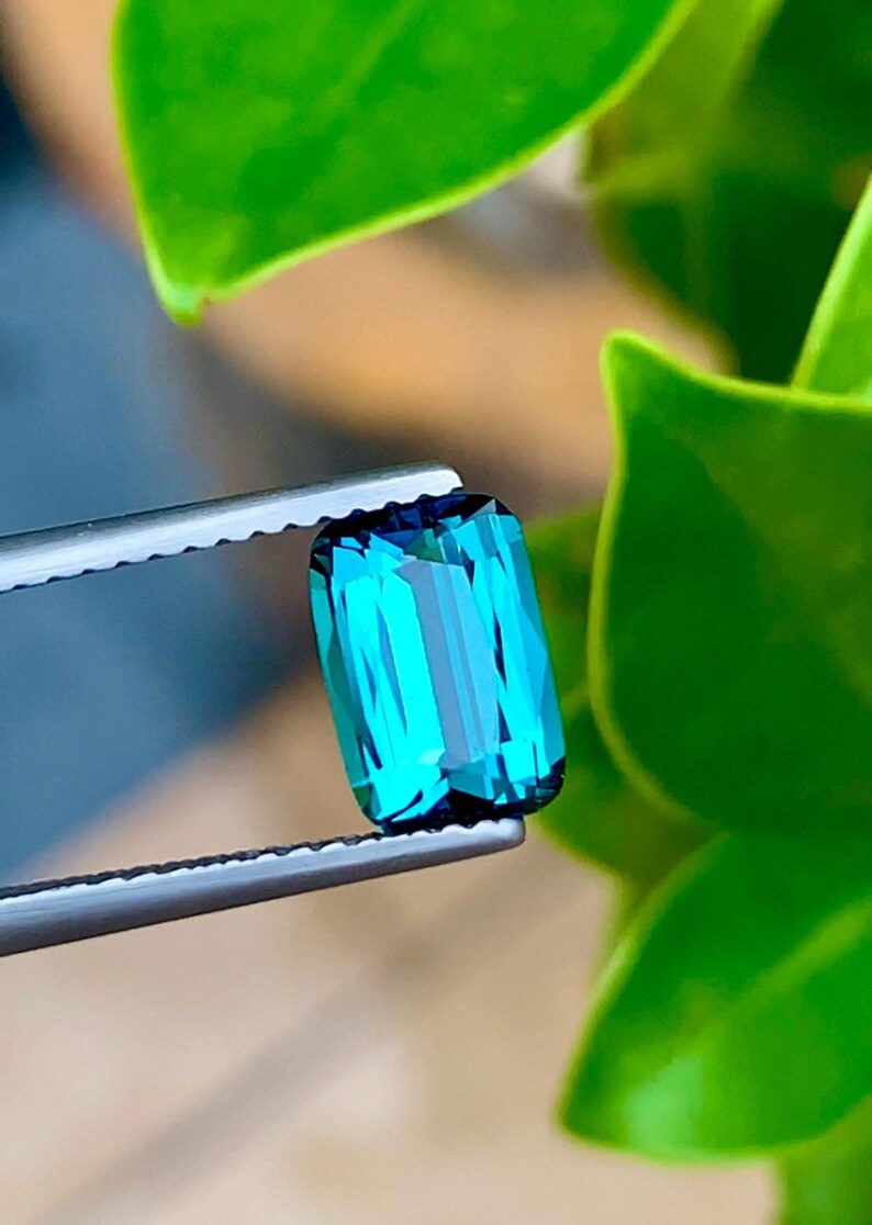 May include: A vibrant teal green gemstone with a rectangular cut, held in tweezers against a backdrop of green foliage.