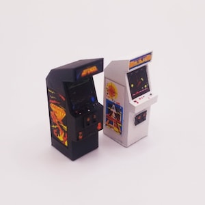 Puede incluir: Dos gabinetes de arcade en miniatura, uno negro y otro blanco, con un diseño de videojuego retro. El gabinete negro tiene un diseño rojo, naranja y amarillo con el texto "GALAXIAN" en la parte superior. El gabinete blanco tiene un diseño azul y amarillo con el texto "GALAXIAN" en la parte superior.