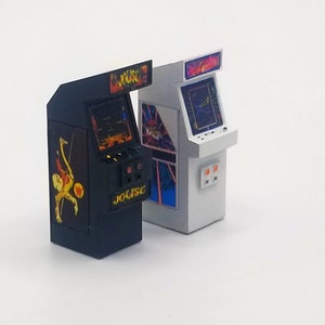 May include: Two miniature arcade cabinets, one black with a Joust graphic and the other white with a Galaga graphic. Both cabinets have a small screen and control buttons.