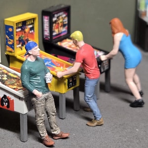 Op de afbeelding: Miniatuurscène met drie flipperkasten met kleurrijke graphics. Figuren van mensen zijn rond de machines geplaatst, wat een speelkameromgeving suggereert. De machines hebben verschillende thema's, waaronder een flipperkast met KISS-thema.