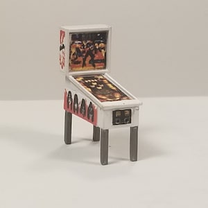 May include: Miniature pinball machine with a white cabinet, red accents, and a detailed playfield. The backboard features a colorful graphic. The machine is supported by four gray legs. A classic arcade game in a small scale.