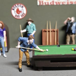 May include: A miniature pool hall scene with several figures. A person is aiming a cue stick at a white ball on a green felt pool table. Other figures are standing around, and there is a jukebox, a sign that says "Budweiser", and a "Boston Red Sox" sign.
