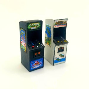 Può includere: Due cabinet arcade in miniatura, uno nero e uno bianco, con il titolo del gioco "Galaxian" su entrambi. Il cabinet nero ha un pulsante rosso e due pulsanti arancioni, mentre il cabinet bianco ha due pulsanti gialli.
