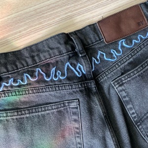 May include: Black denim jeans with a blue wavy stitch detail around the waistband. A brown leather patch with the letters "RV" is sewn on the back right side of the waistband.