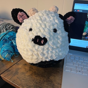 May include: A white and black crocheted plush toy shaped like a cow with pink ears and a black nose. The toy is sitting on a wooden table next to a laptop.