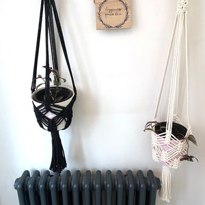 May include: Two macrame plant hangers, one black and one white, hanging from hooks on a white wall. The black hanger has a white pot with a pink and white pattern and a small green plant. The white hanger has a pink pot with a white pattern and a small green plant. A wooden sign with the text "Happiness grows here" hangs between the two plant hangers.