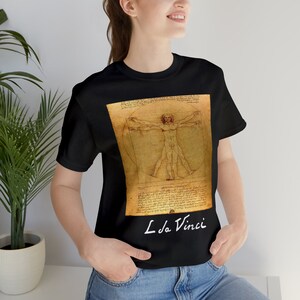 May include: A black t-shirt featuring a graphic print of Leonardo da Vinci's Vitruvian Man drawing on a tan parchment-colored background. The design includes the iconic anatomical figure within a circle and square, accompanied by handwritten text and the signature L. da Vinci in white at the bottom.