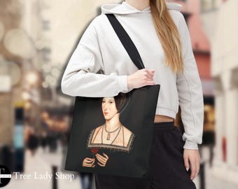 Pode incluir: Bolsa tote preta com um retrato de uma mulher em trajes históricos, segurando uma rosa vermelha. O retrato tem um fundo preto. A bolsa tem uma alça preta e está sendo carregada por uma pessoa.