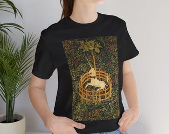 Pode incluir: T-shirt preto com um design de tapeçaria que apresenta um unicórnio dentro de uma cerca de madeira, com um fundo de árvores e folhagens. A obra de arte tem um estilo medieval, com um fundo floral detalhado.