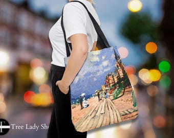 Pode incluir: Bolsa tote com uma pintura de uma cena costeira, com um céu azul e figuras numa passarela de madeira. A bolsa tem uma alça preta e é usada ao ombro. A obra de arte retrata edifícios e uma praia.