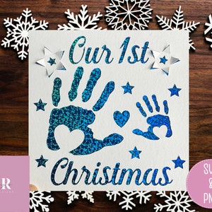 May include: A white card with blue glitter handprints and the text "Our 1st Christmas". The card is surrounded by white snowflake cutouts.