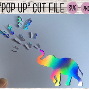 Puede incluir: Un elefante iridiscente arcoíris con mariposas que vuelan de su trompa. El elefante y las mariposas están recortados en papel y forman parte de una tarjeta pop-up.