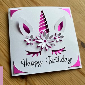 May include: A white card with a pink unicorn design and the text "Happy Birthday". The unicorn has a pink horn, pink ears, and pink eyelashes. There are white flowers around the unicorn's horn.