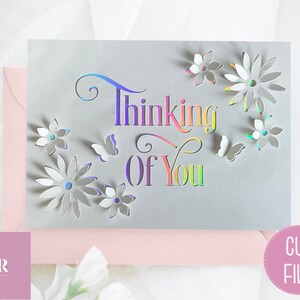 May include: A gray card with the words "Thinking of You" in rainbow colors and cut-out flowers and butterflies. The card is on a pink surface.