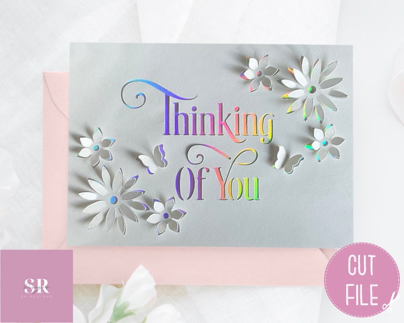 May include: A gray card with the words "Thinking of You" in rainbow colors and cut-out flowers and butterflies. The card is on a pink surface.