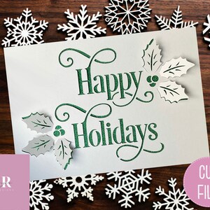 Puede incluir: Una tarjeta blanca con letras verdes brillantes que dicen "Felices Fiestas" con hojas de acebo y bayas a cada lado. La tarjeta est&aacute; rodeada de copos de nieve blancos sobre un fondo de madera.