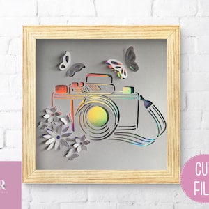 May include: A paper cut file design of a camera with a strap, flowers, and butterflies. The design is in a wooden frame with a gray background.