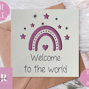 May include: A white card with a pink glitter rainbow and stars. The text "Welcome to the world" is written below the rainbow.