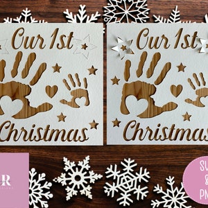May include: Two white cards with the text "Our 1st Christmas" and a handprint design. The cards are on a wooden surface with white snowflakes.