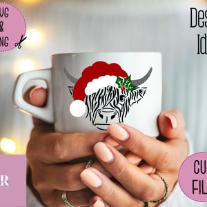 K&ouml;nnte beinhalten: Eine wei&szlig;e Keramiktasse mit einer schwarz-wei&szlig;en Highland-Kuh, die einen roten Weihnachtsmann-Hut und einen gr&uuml;nen Stechpalmenzweig tr&auml;gt. Die Tasse wird von den H&auml;nden einer Person gehalten.