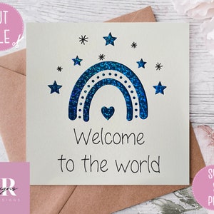 May include: A white card with a blue glitter rainbow and stars. The text "Welcome to the world" is written below the rainbow.