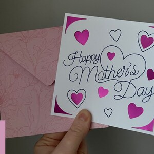 May include: A white card with the text "Happy Mother's Day" in black cursive script. The card is decorated with pink hearts and a pink envelope with a floral pattern.