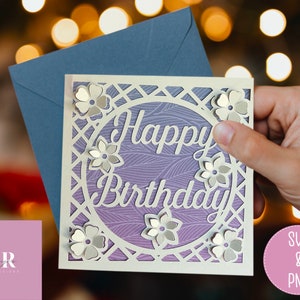 Op de afbeelding: Een witte kaart met een paarse achtergrond en een wit tralieontwerp. De kaart heeft een cirkel in het midden met de woorden "Happy Birthday" in wit handschrift. De cirkel is omringd door witte bloemen.