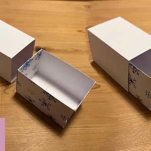 May include: A white box with a blue and gold snowflake pattern is partially open, revealing a white interior. The box is on a wooden surface. The text "CUT FILE" and "SVG" are visible in the image.